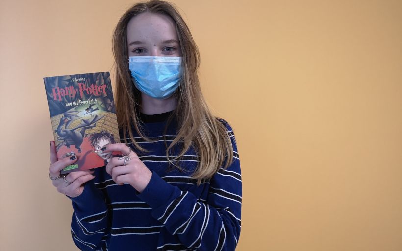 Eine Schülerin der 9. Klasse steht mit einem Buch in der Hand vor einer orangefarbenen Wand.