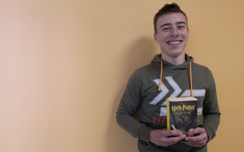 Ein Schüler der 9. Klasse steht mit einem Buch in der Hand vor einer orangefarbenen Wand.