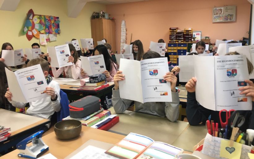 Schüler sitzen in einem Klassenzimmer und halten sich eine Zeitung vor das Gesicht.