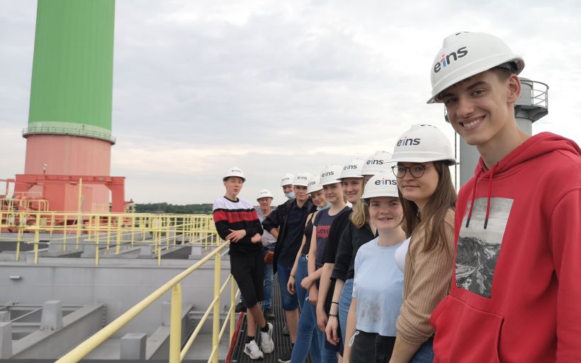 Schüler stehen mit Helmen auf dem Kopf auf dem Dach eines Hezkraftwerks neben einem bunten Schonstein.
