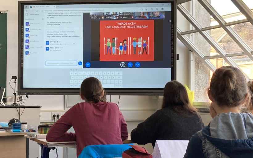 Schüler sitzen in einem Klassenzimmer und blicken auf eine Videokonferenz an einer digitalen Tafel.