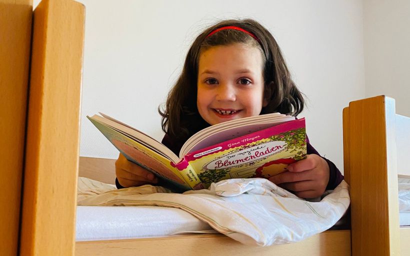Ein lächelndes Mädchen schaut aus ihrem Hochbett und hält ein Lesebuch in der Hand.